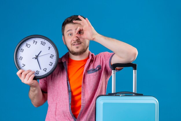 Hombre joven guapo viajero sosteniendo reloj y maleta sonriendo positivo