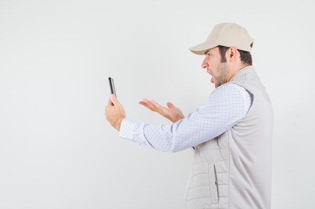 Hombre joven hablando con alguien a través de una videollamada con chaqueta y gorra beige y mirando furioso, vista frontal.