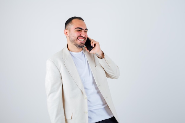 Hombre joven hablando por teléfono en camiseta blanca, chaqueta y mirando feliz. vista frontal.