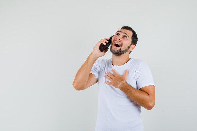 Hombre joven hablando por teléfono en camiseta blanca y mirando avergonzado, vista frontal. espacio para texto