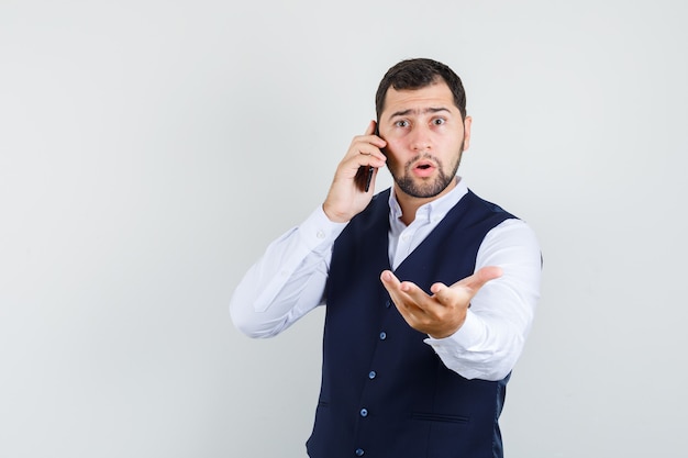 Hombre joven hablando por teléfono celular con la mano levantada en camisa y chaleco y mirando desconcertado