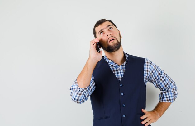 Hombre joven hablando por teléfono móvil en camisa, chaleco y mirando pensativo.
