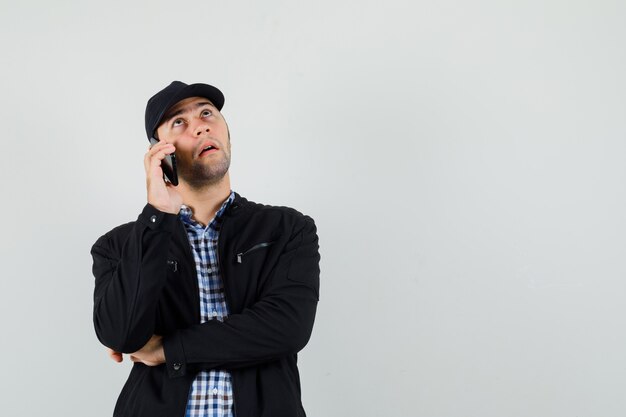 Hombre joven hablando por teléfono móvil en camisa, chaqueta, gorra y mirando pensativo, vista frontal.