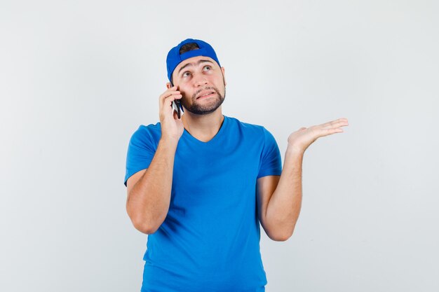 Hombre joven hablando por teléfono móvil en camiseta azul y gorra y mirando indefenso