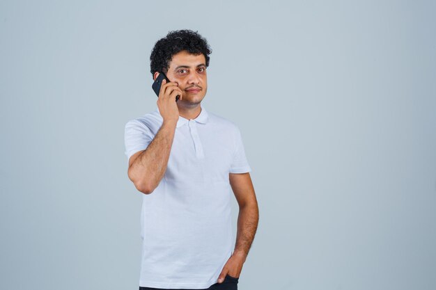 Hombre joven hablando por teléfono móvil en camiseta blanca y mirando esperanzado, vista frontal.