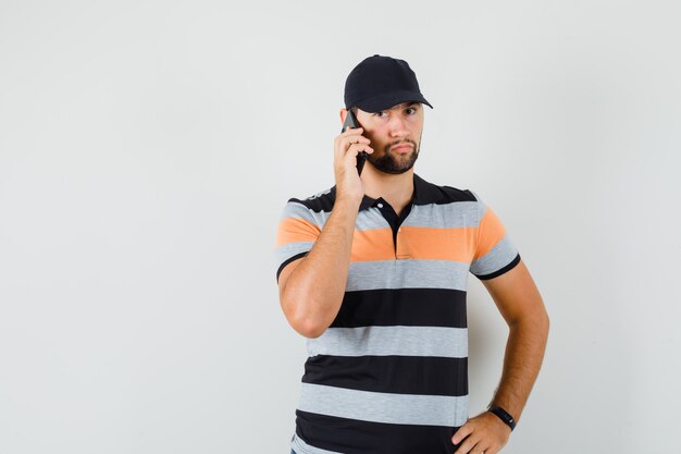 Hombre joven hablando por teléfono móvil en camiseta, gorra y mirando serio.