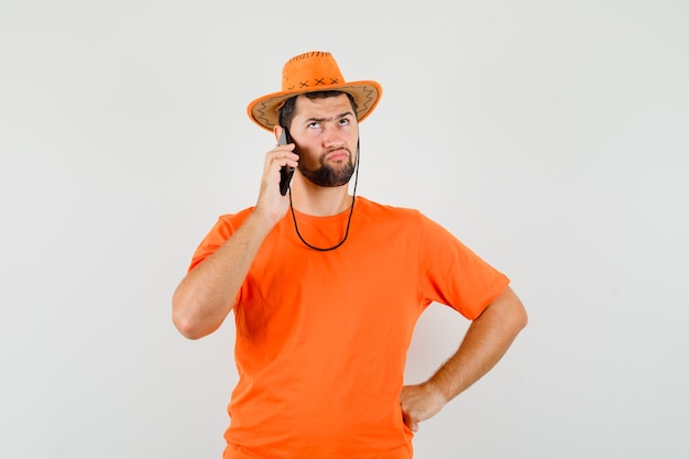 Hombre joven hablando por teléfono móvil en camiseta naranja, sombrero y mirando indeciso, vista frontal.