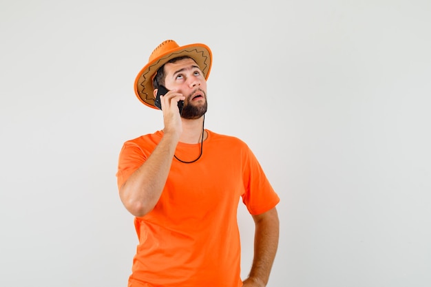 Hombre joven hablando por teléfono móvil en camiseta naranja, sombrero y mirando pensativo. vista frontal.