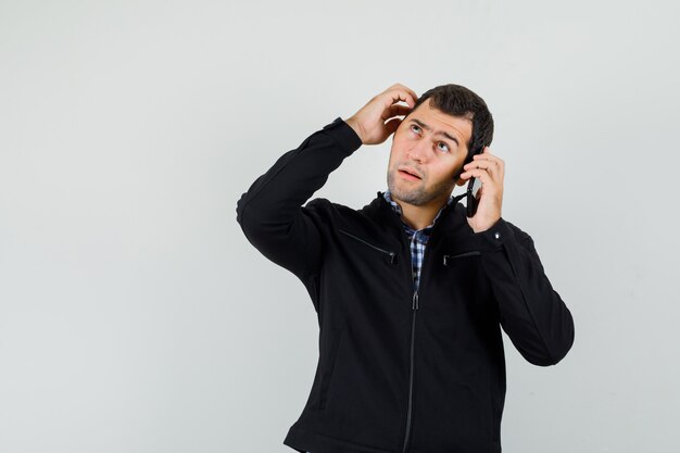 Hombre joven hablando por teléfono móvil, rascándose la cabeza en camisa, chaqueta y mirando pensativo. vista frontal.