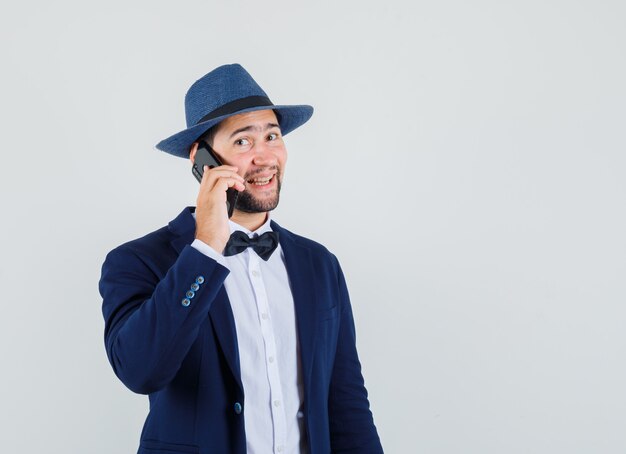 Hombre joven hablando por teléfono móvil en traje, sombrero y mirando contento, vista frontal.