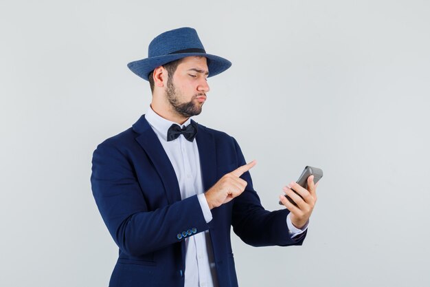 Hombre joven haciendo cálculos en la calculadora en traje, sombrero y mirando enfocado. vista frontal.