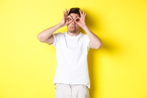 Hombre joven haciendo muecas y mostrando la lengua, tonto, de pie en camiseta blanca sobre fondo amarillo. Copia espacio