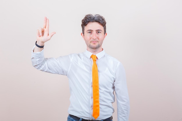 Hombre joven haciendo signo de pistola de dedo en camisa blanca, corbata y mirando confiado