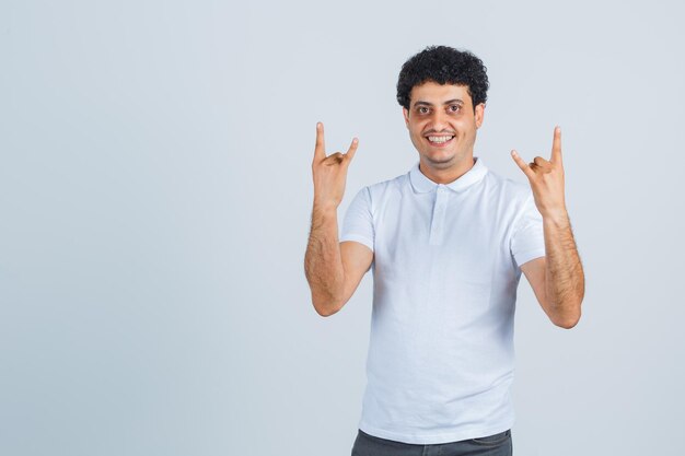 Hombre joven haciendo símbolo de rock en camiseta blanca, pantalón y mirando dichoso. vista frontal.