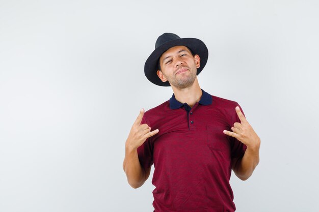 Hombre joven haciendo símbolo de rock en camiseta, sombrero y luciendo genial. vista frontal.