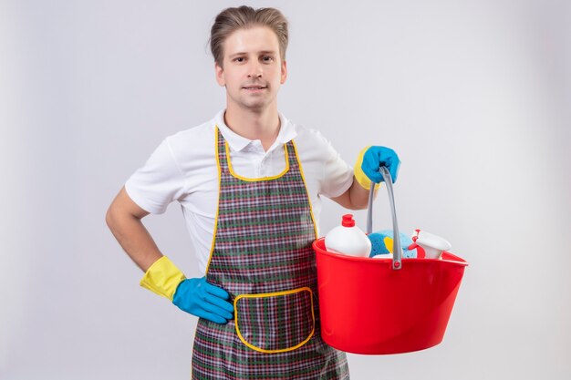 Hombre joven hansdome con delantal y guantes de goma sosteniendo un balde con herramientas de limpieza con sonrisa segura