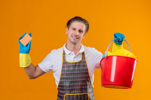 Hombre joven hansdome con delantal y guantes de goma sosteniendo un balde con herramientas de limpieza