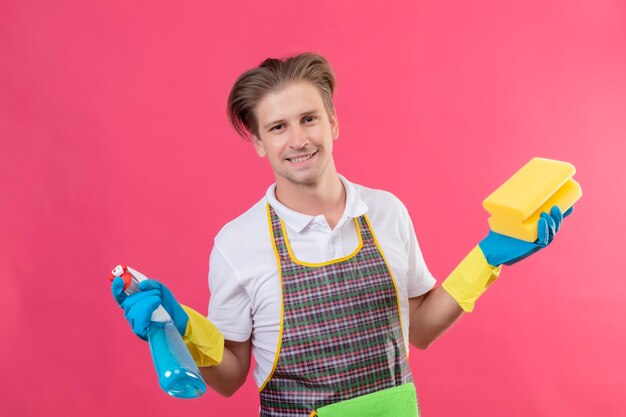 Hombre joven hansdome con delantal y guantes de goma sosteniendo spray de limpieza y esponja con una gran sonrisa en la cara feliz y positiva de pie sobre la pared rosa