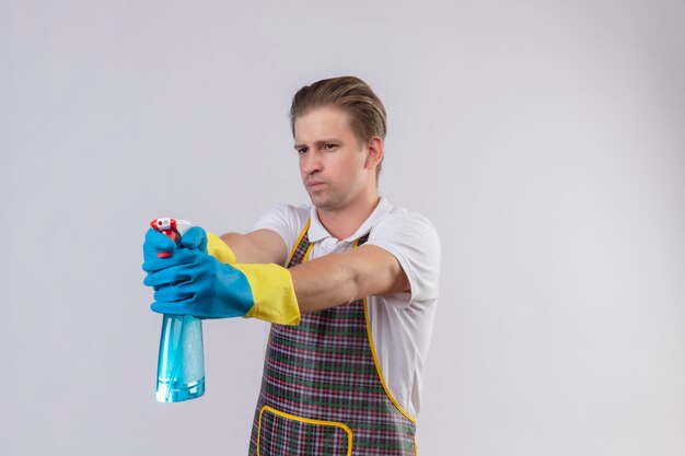 Hombre joven hansdome con delantal y guantes de goma sosteniendo spray de limpieza usando como una pistola con cara seria de pie sobre una pared blanca