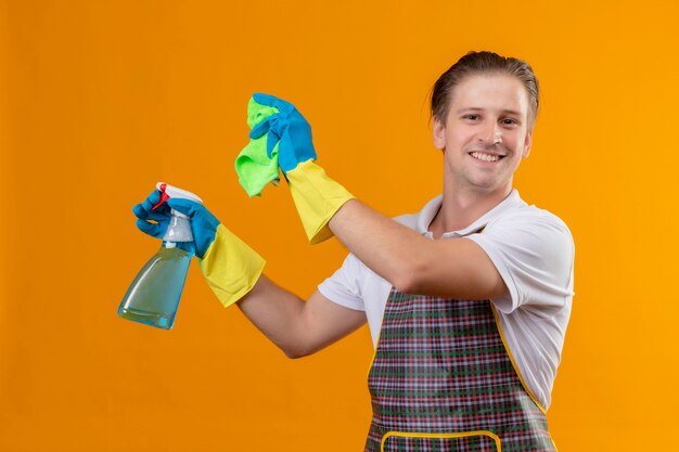 Hombre joven de Hansdome con delantal sosteniendo spray de limpieza y alfombra feliz y positivo sonriendo listo para limpiar de pie sobre la pared naranja