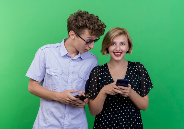 Hombre joven hermosa pareja espiando mirando el teléfono celular de su novia de pie sobre la pared verde