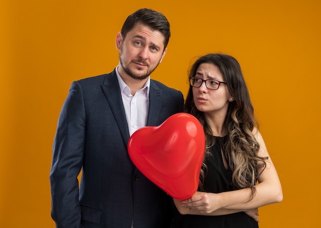 Hombre joven y hermosa pareja y mujer resentida con globo rojo en forma de corazón de pie uno al lado del otro celebrando el día de San Valentín