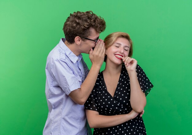 Hombre joven hermosa pareja susurrando un secreto a su novia sonriente de pie sobre la pared verde