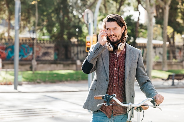 Foto gratuita hombre joven hermoso con el auricular alrededor de su cuello que habla sobre el teléfono móvil en el camino