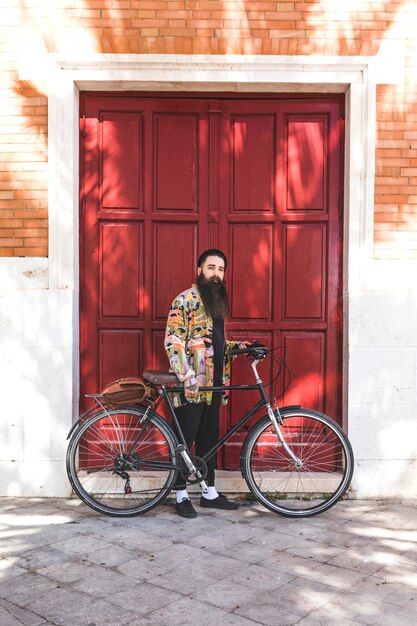 Hombre joven hermoso con la bicicleta que se coloca delante de la pared de madera de la puerta roja