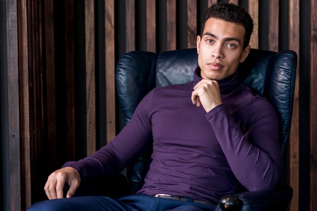 Hombre joven hermoso en la camiseta del cuello del polo que se sienta en la butaca que mira la cámara