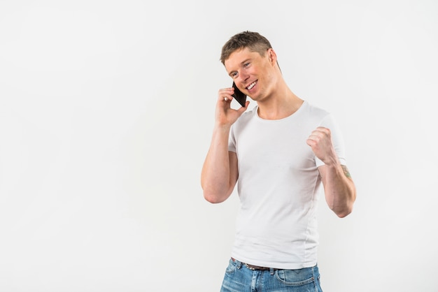 Hombre joven hermoso feliz que aprieta su puño que habla en el teléfono móvil contra el fondo blanco