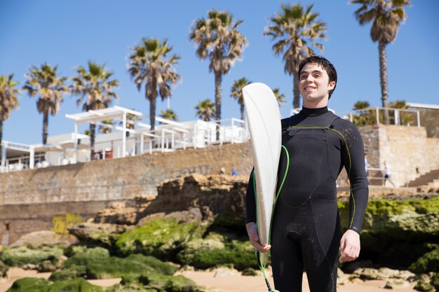 Hombre joven hermoso feliz que sostiene la tabla hawaiana en la playa soleada