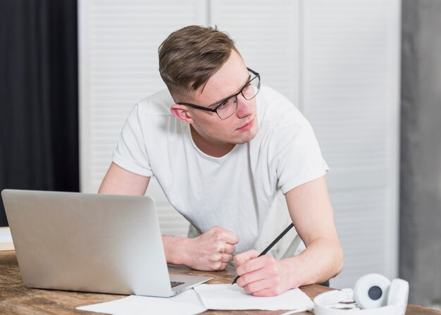 Hombre joven hermoso que mira la escritura ausente en el papel con el lápiz y el ordenador portátil en la tabla