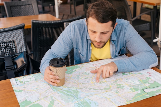 Hombre joven hermoso que mira el mapa con sostener la taza de café en caf�