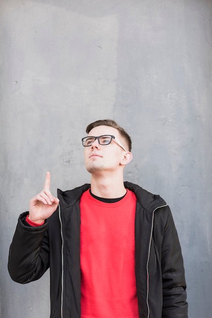 Foto gratuita hombre joven hermoso que señala su dedo hacia arriba que mira para arriba contra el muro de cemento