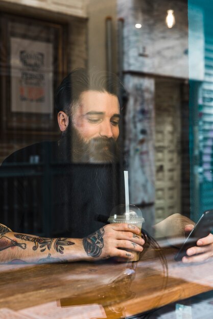 Hombre joven hermoso que se sienta en café usando el teléfono móvil