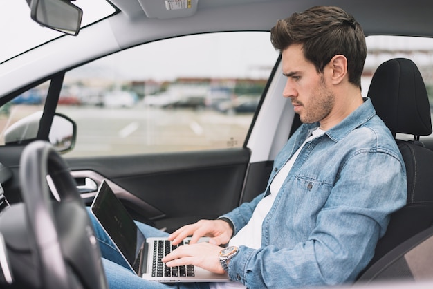Hombre joven hermoso que se sienta dentro del coche usando el ordenador portátil