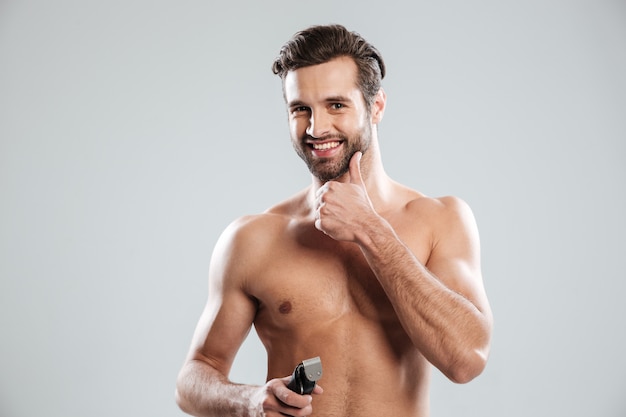 Hombre joven hermoso que sostiene la maquinilla de afeitar eléctrica y tocando su barbilla