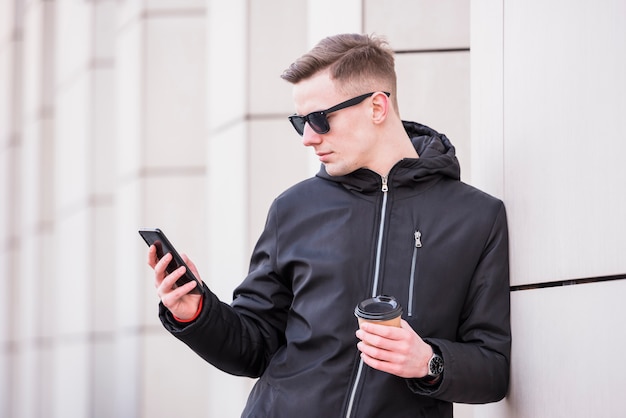 Foto gratuita hombre joven hermoso que sostiene la taza de café para llevar usando smartphone