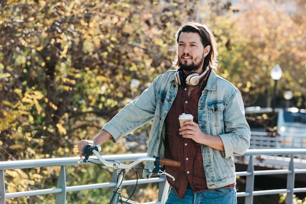 Hombre joven hermoso que sostiene la taza de café de papel que se coloca con la bicicleta en el parque