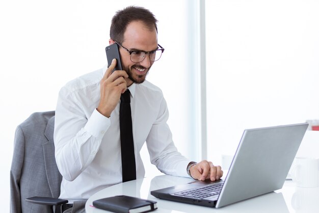 Hombre joven hermoso que trabaja con la computadora portátil y el teléfono móvil en la oficina.