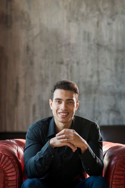 Hombre joven hermoso sonriente que se sienta en la butaca contra la pared gris