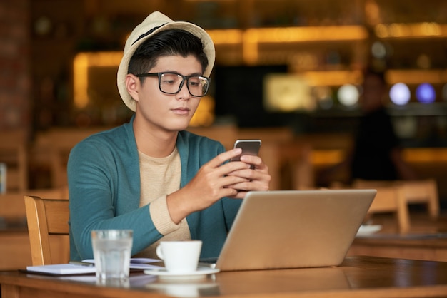 Hombre joven inconformista asiático sentado en la cafetería con ordenador portátil y con teléfono inteligente