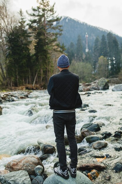 Hombre joven inconformista caminando sobre una roca en el río en el bosque de invierno