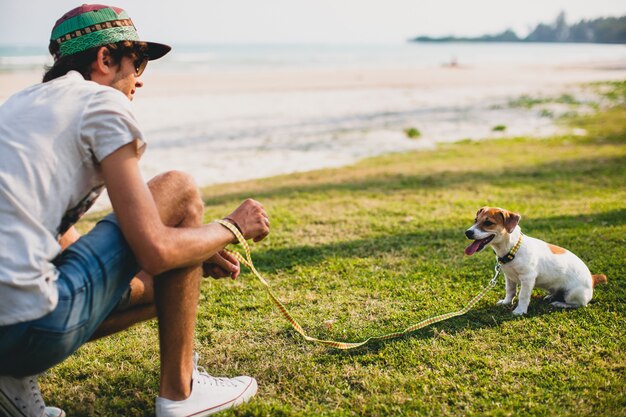 Hombre joven inconformista con estilo caminando y jugando con perro en playa tropical