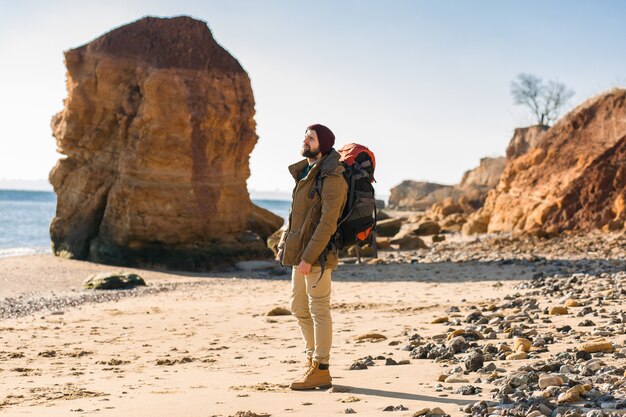 Hombre joven inconformista que viaja solo con mochila en la costa del mar de otoño vistiendo chaqueta y sombrero