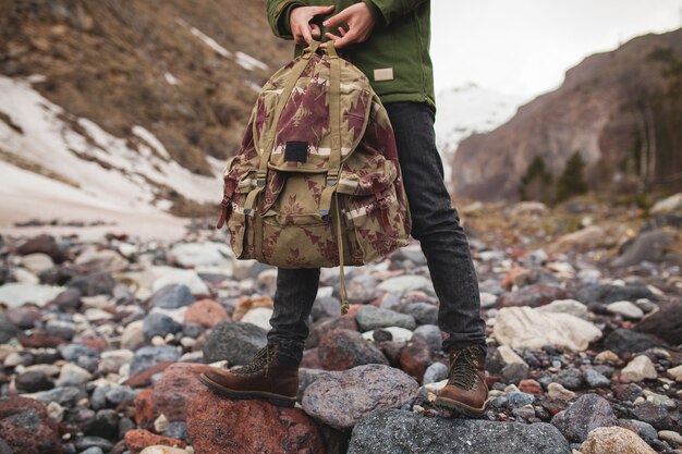 Hombre joven inconformista, senderismo por el río, naturaleza salvaje, vacaciones de invierno, sosteniendo la mochila en las manos, detalles de cerca