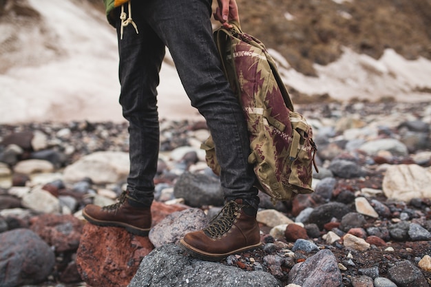 Hombre joven inconformista, senderismo por el río, naturaleza salvaje, vacaciones de invierno, sosteniendo la mochila en las manos, detalles de cerca