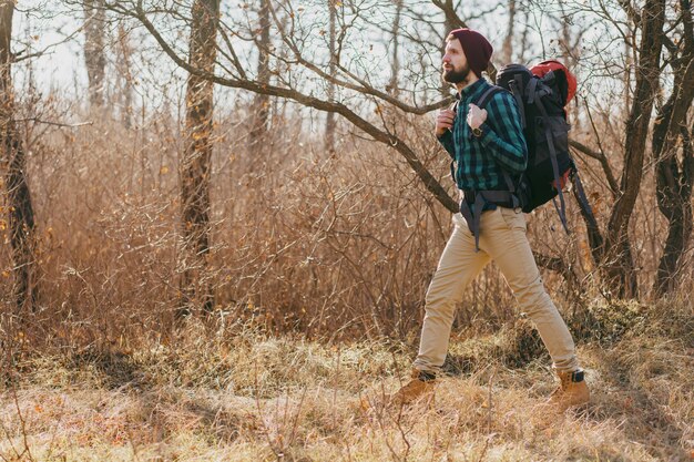 Hombre joven inconformista viajando con mochila en el bosque de otoño con camisa a cuadros y sombrero, turista activo caminando, explorando la naturaleza en la estación fría