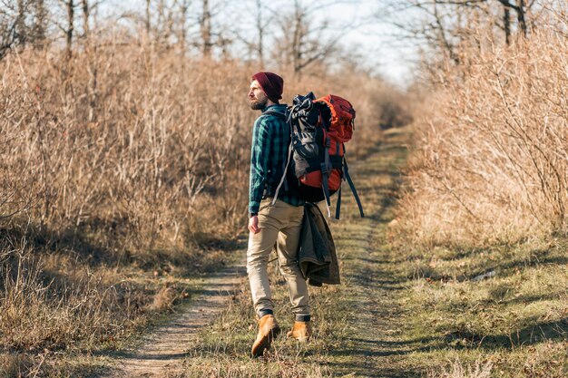 Hombre joven inconformista viajando con mochila en el bosque de otoño con camisa a cuadros y sombrero, turista activo corriendo, explorando la naturaleza en la estación fría